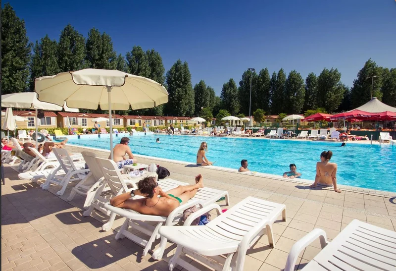 People lounging on sunbeds and swimming in a large pool, surrounded by trees and holiday chalets at hu Venezia Camping in Town