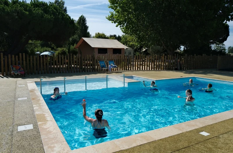 People swimming in a clear blue pool surrounded by trees and wooden fencing, with tents and lounge chairs nearby at Seasonova Ile de Ré