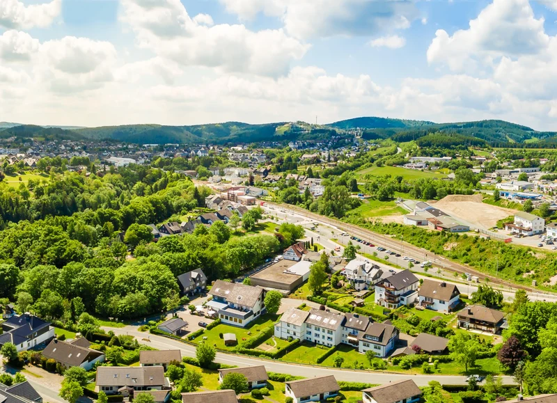 Aerial view: Small town surrounded by lush green hills and a railway, under partly cloudy skies at Bergresort Winterberg