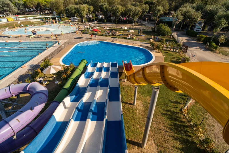 Water slides descend into pools, surrounded by sunbathers and umbrellas; trees and paths frame the scene at Marina Julia Family Camping Village