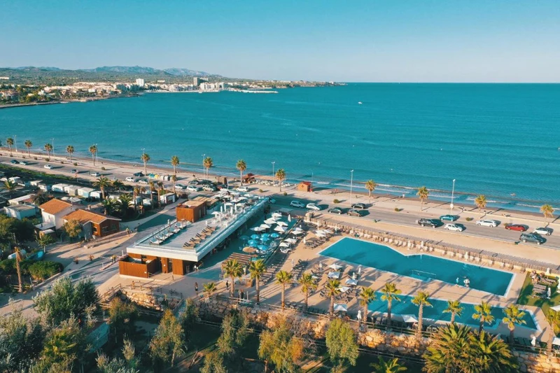 Large pool surrounded by deck chairs and umbrellas, facing a beach road with palm trees and ocean at Taïga Delta De L'Ebre