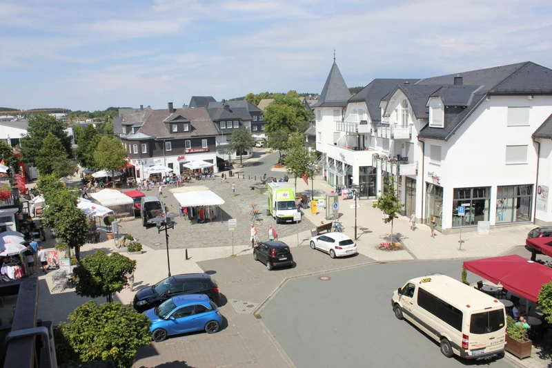 Market square with outdoor tents and parked cars amid shops and cafes, surrounded by residential buildings at Bergresort Winterberg