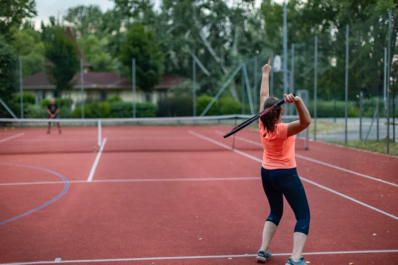 Person prepares to serve in tennis game on an outdoor red court with trees and buildings in the background at Füred