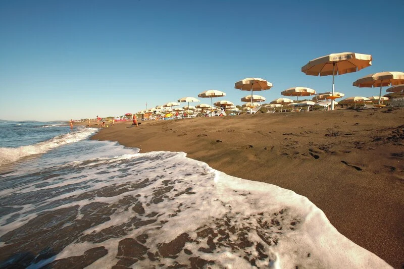 Waves gently wash onto a sandy beach lined with numerous tan umbrellas, set against a clear blue sky at Club Degli Amici