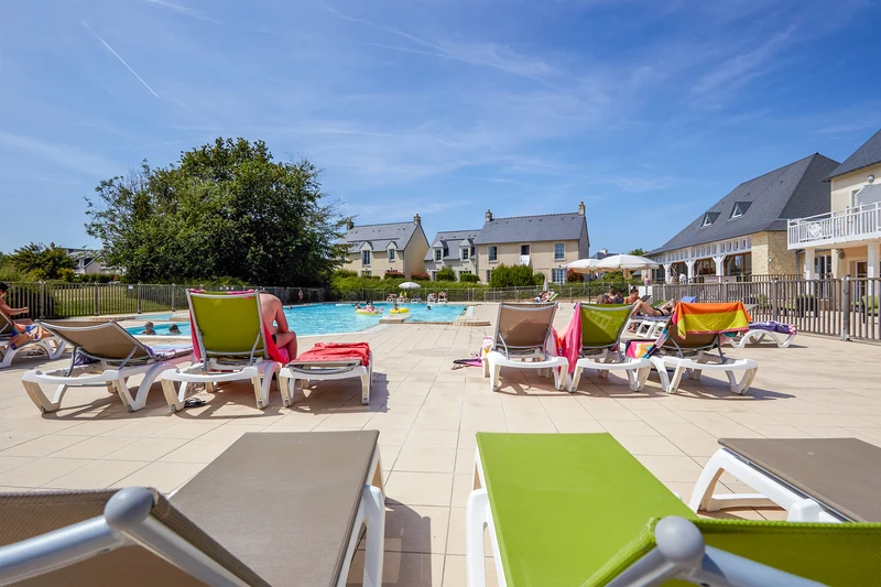 Sunbathers relax on loungers beside a clear swimming pool, surrounded by residential cottages under a bright blue sky at Le Green Beach