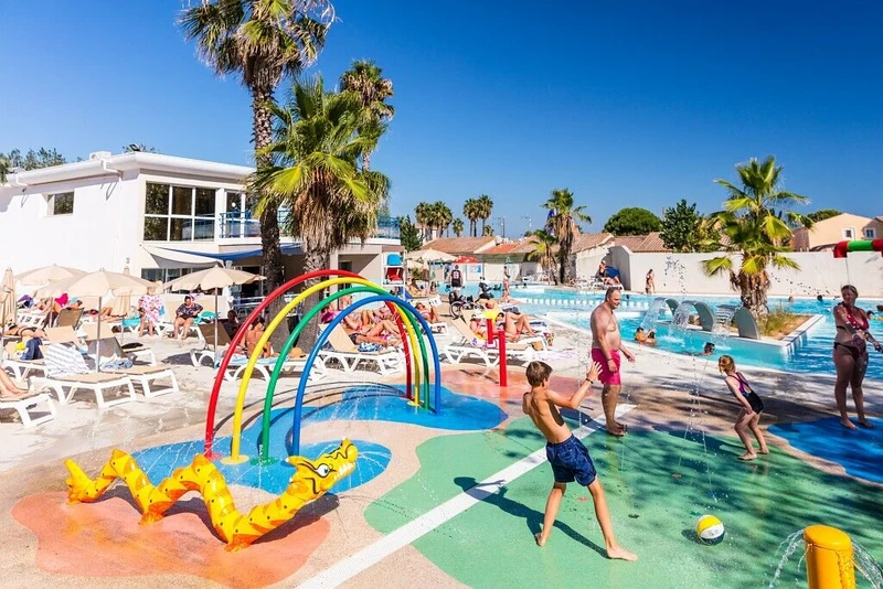 Children play in a splash pad with colorful arches beside a pool, surrounded by lounging adults and palm trees at Marvilla Parks Club Le Napoléon