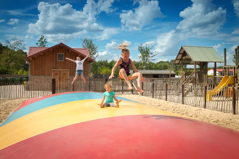 Three children bouncing on an inflatable play structure in a sandy playground with wooden buildings in the background at 't Geuldal