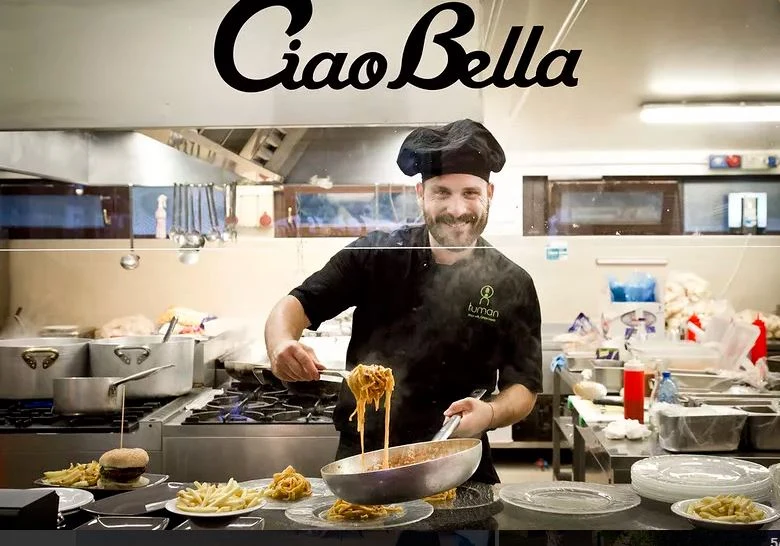 Chef cooking pasta in a busy kitchen with various dishes around, smiling behind a glass window at hu Venezia Camping in Town