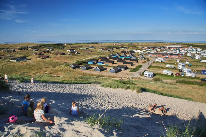 Children play on sand dunes, overlooking a sprawling campsite with cabins and trailers under a clear blue sky at Dancamps Nordsø