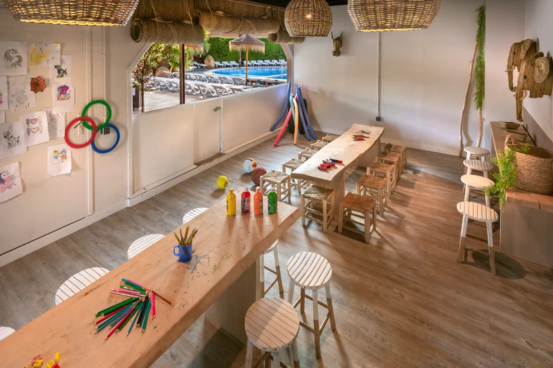 Wooden tables arranged for a kids' craft session with drawings on wall; large windows reveal a sunny pool area at Enmar