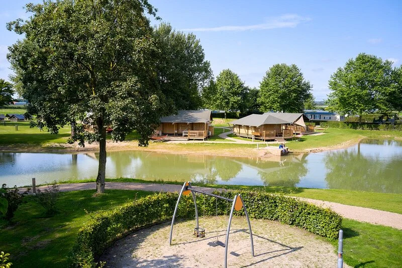 Wooden cabins by a pond, surrounded by trees, with a swing set in the foreground at Betuwe
