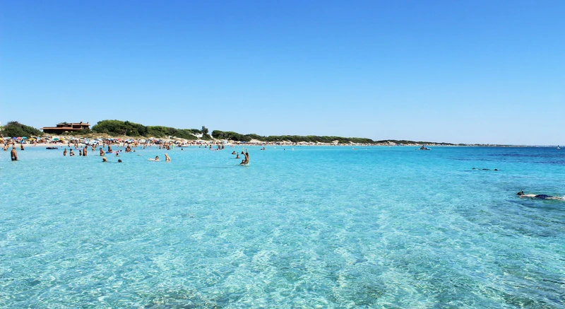 Swimmers enjoying clear, blue water on a sunny day; crowded beach with sunbathers in the background at Porto Cesareo Camping