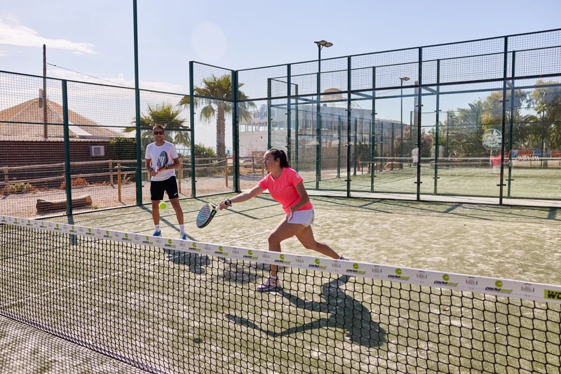 Two people play paddle tennis on a fenced outdoor court in sunny weather, near palm trees and a building at Taïga Delta De L'Ebre