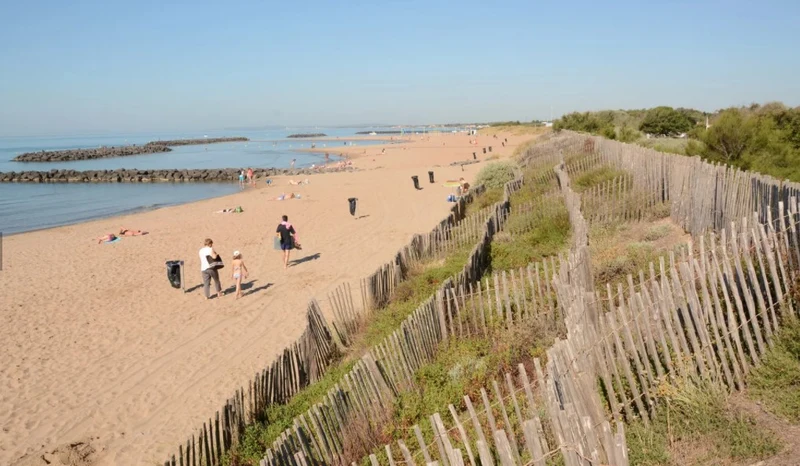 People walking and relaxing on a serene sandy beach beside calm sea waters and dune fences at Marvilla Parks Club Le Napoléon