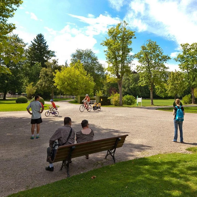 Two cyclists and child riding bikes on a gravel path, observed by people sitting and standing, surrounded by trees at Seasonova Vittel