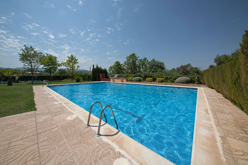 Swimming pool reflecting clear blue sky, surrounded by trees and hedges, at a tranquil campsite at Els Ports