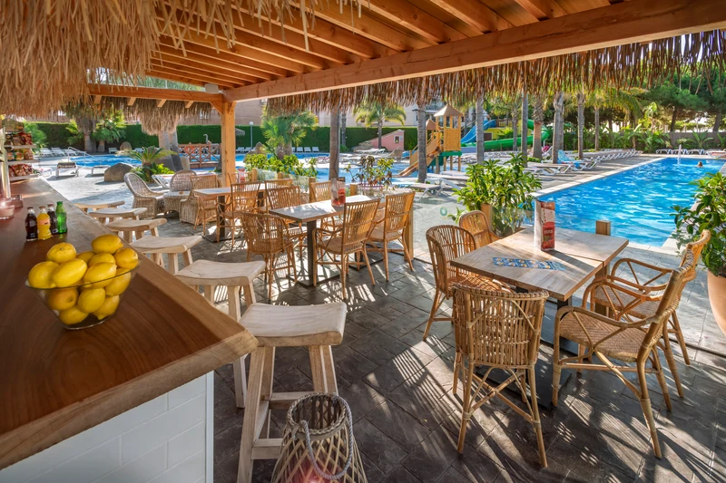 Bar with stools and tables under wooden canopy overlooking pool area with lounge chairs and greenery at Enmar
