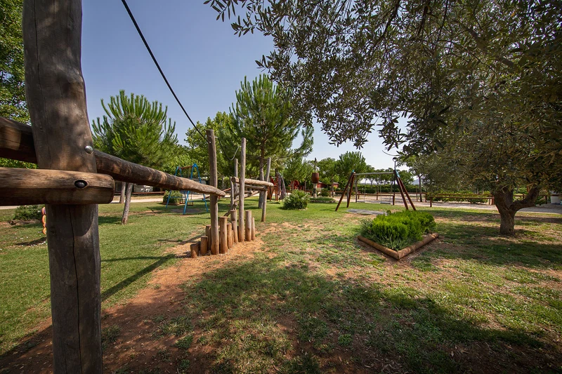 Wooden playground equipment stands on a grassy area surrounded by trees and other play structures at Els Ports