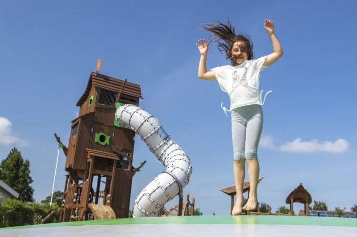 Child jumping joyfully on a trampoline near a wooden play structure with slides and climbing features at Camping de Molenhoek